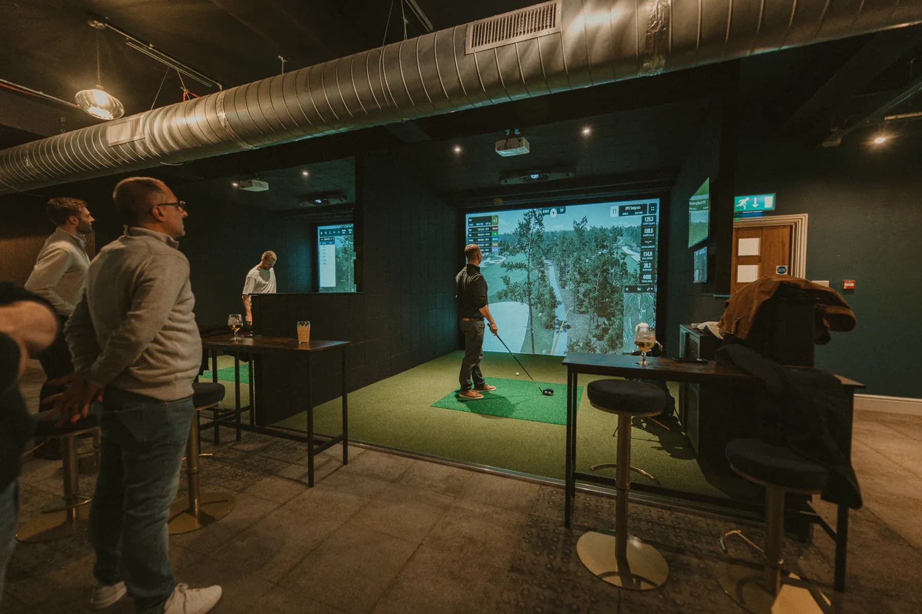 Two simulator bays side by side, a player teeing off while friends watch with drinks