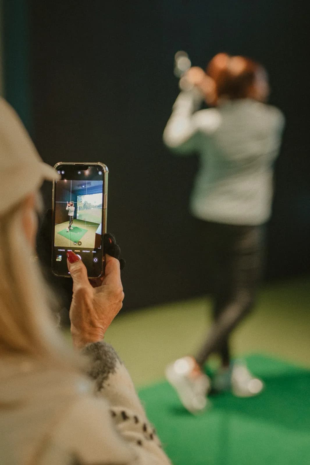 A coach filming a golfer's swing on a phone in a SKRATCH simulator bay