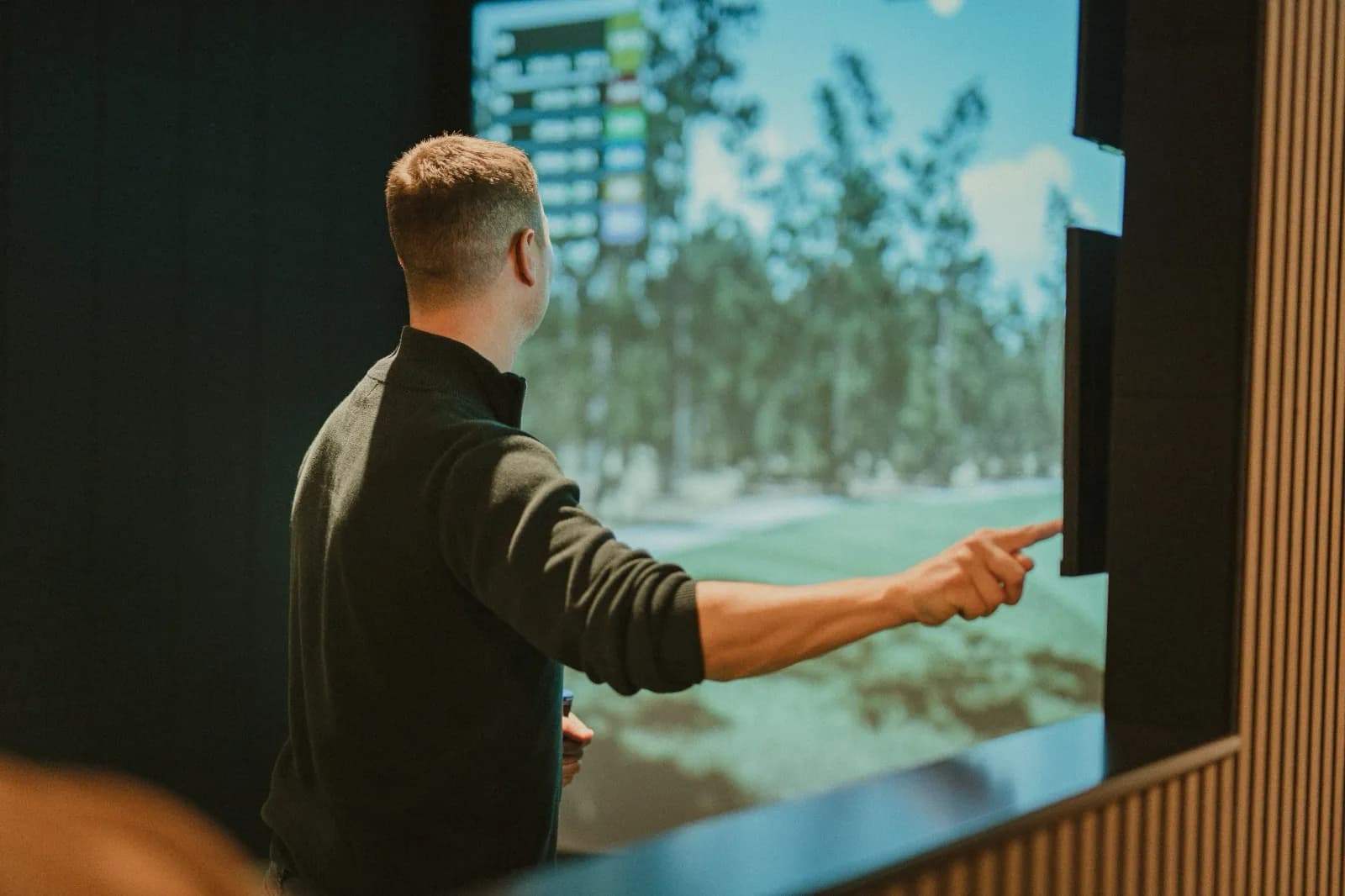 A coach pointing at the simulator screen in a SKRATCH bay, a tree-lined course on the display