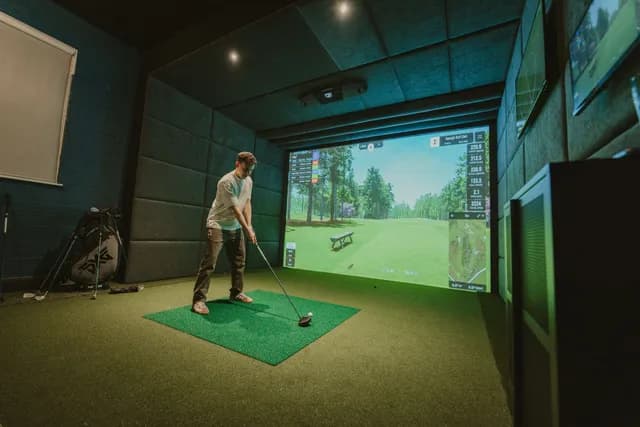 A golfer teeing off in a simulator bay, a tree-lined fairway on screen