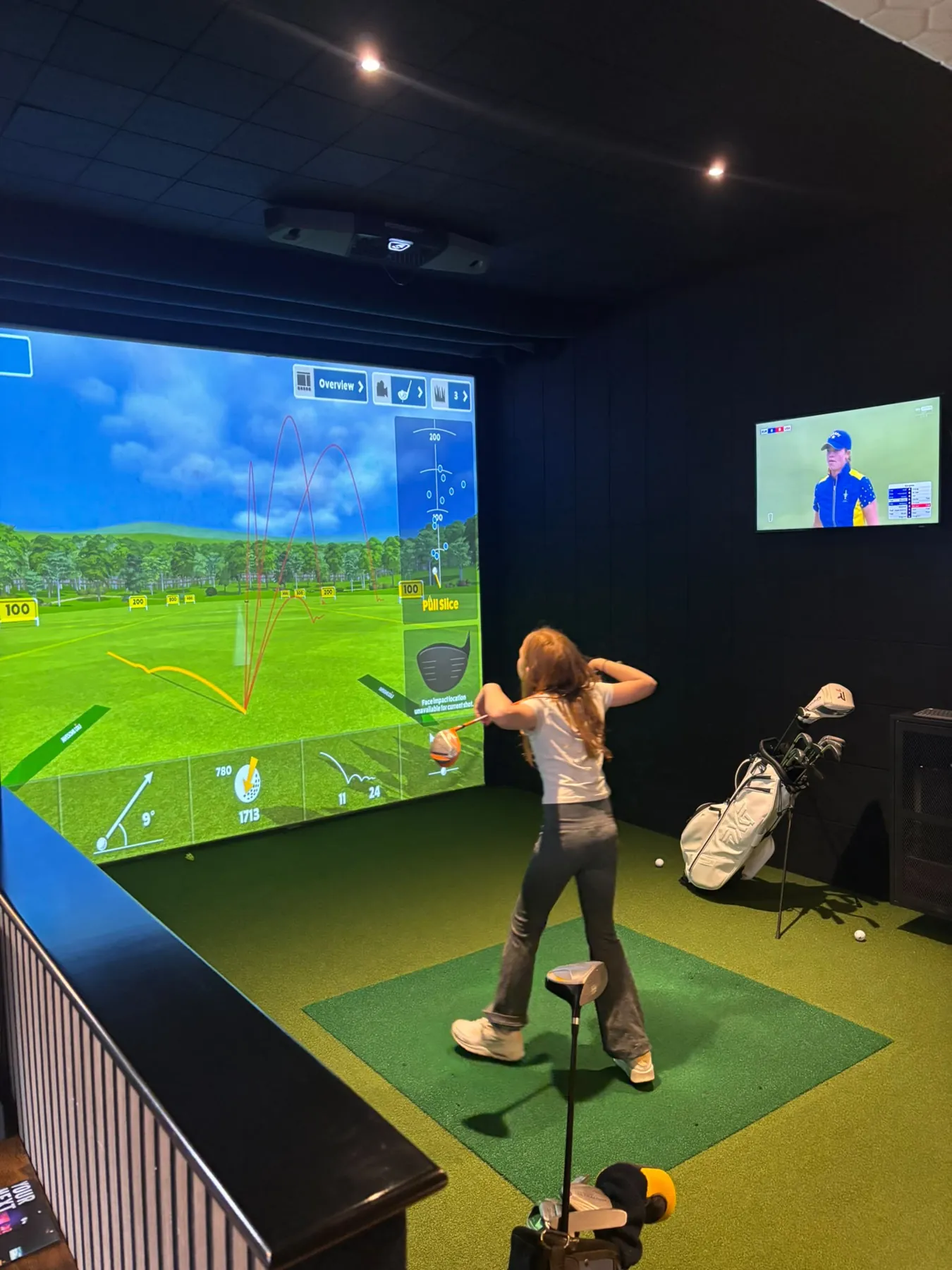 A young golfer follows through on a drive, her shot traced on the simulator screen